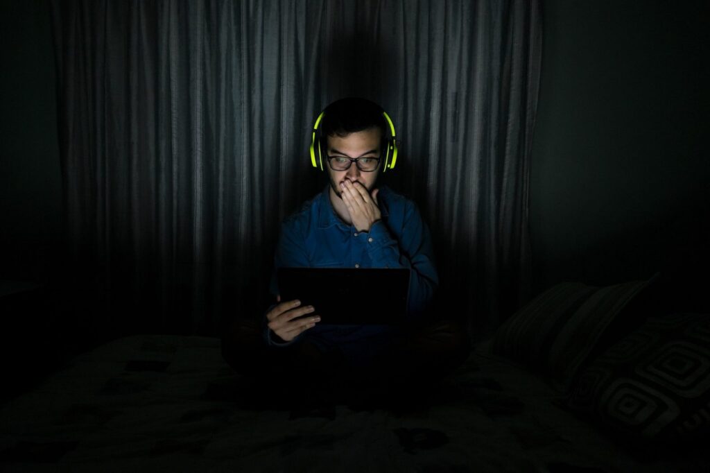 A man in headphones watches a tablet in a dark room, hand over his mouth